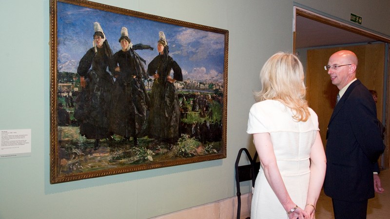 Two people viewing an impressionist painting showing three identically dressed people in old-fashioned black dresses and white hats