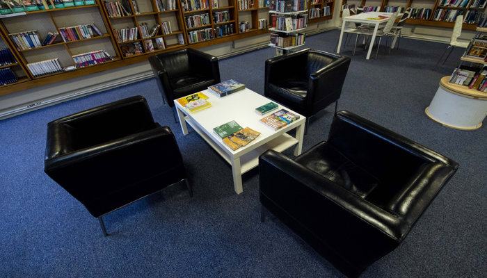 An inside view of Bailliston Library. The library has dark brown bookshelves and a blue carpet. There are 4 black chairs surrounding a white table.