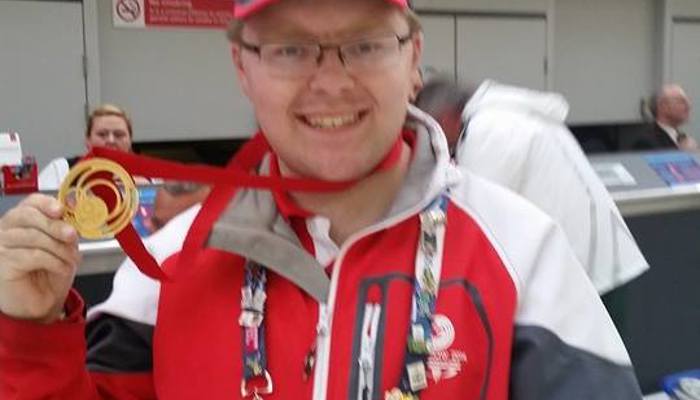 Andrew McVie wearing his red, white and grey Clyde-sider uniform in Glasgow Airport, and he has one of the Glasgow 2014 gold medals around his neck.