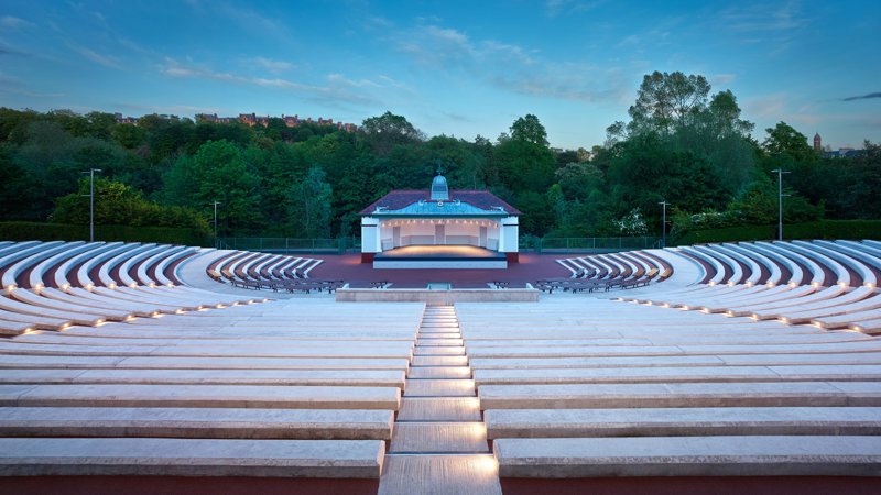 An empty Kelvingrove Bandstand with tall green trees in the background and curved concrete seating, pictured in evening light