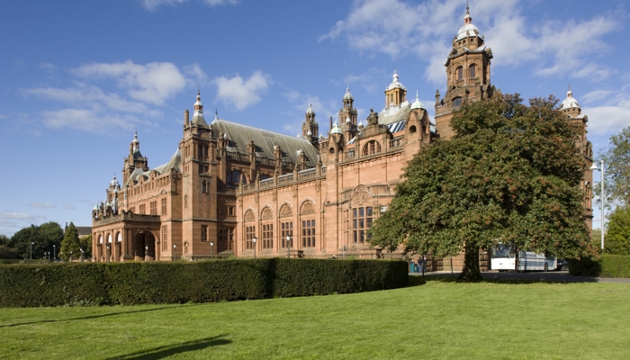 External view of the front of Kelvingrove Art Gallery & Museum with a bright blue sky in the background with a tree and grass lawn to the front of the building