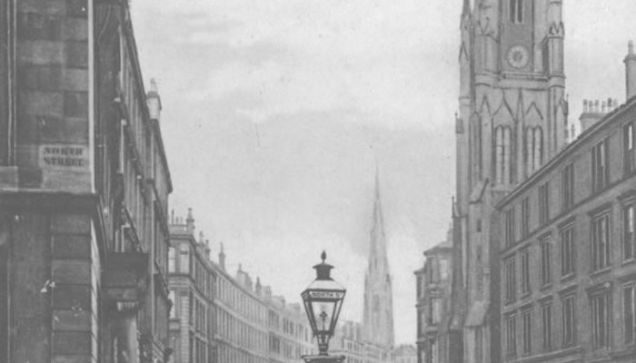 A black and white photo of a street corner on a quiet road with few people and horse and carriages, with tenement buildings and cobbled road.