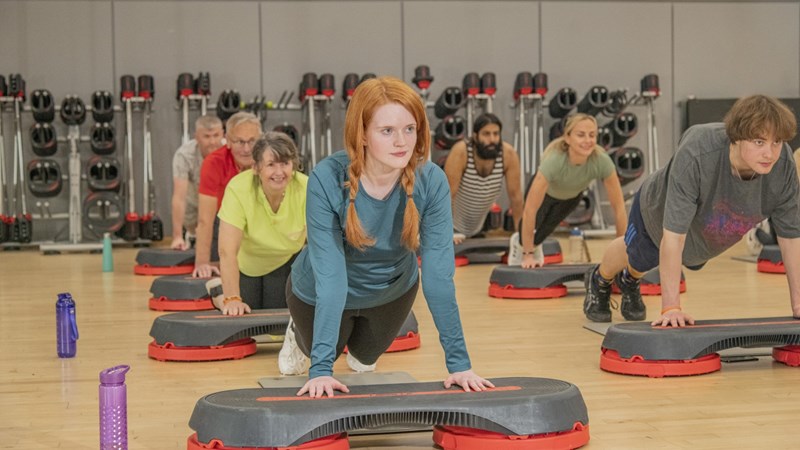 A mix of people in a gym class leaning on a step doing a press up. There is gym equipment in the background.
