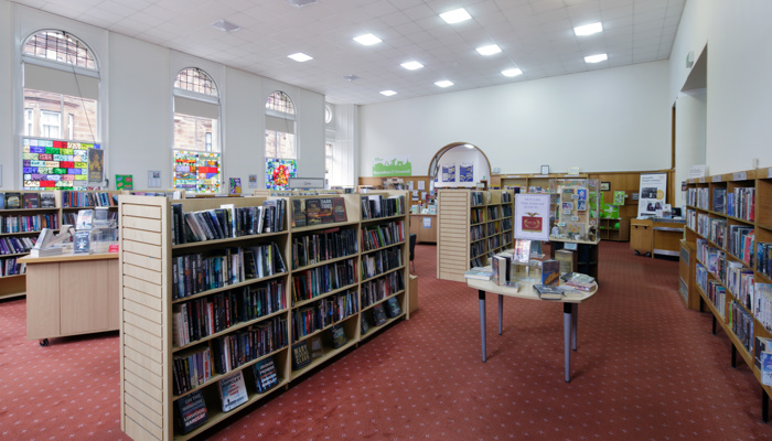 A full bookcase in a library. The floor has red carpet and there are white walls and tall windows in the background.