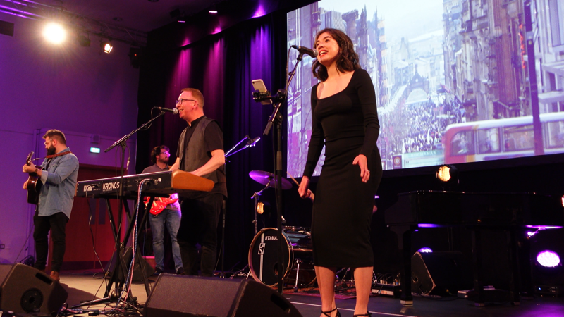 4 people on a stage, 2 of them singing whilst one also plays keyboard and the other 2 playing guitars. There is spotlights shining on the stage and a large bright screen in the background