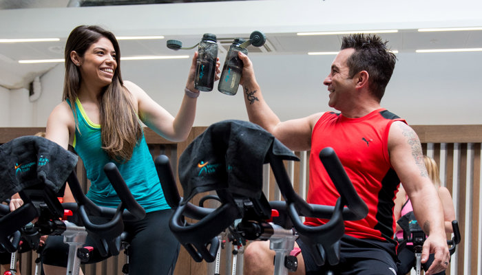 man and woman on spin bikes holding up their water bottles and smiling 