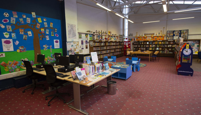 Main view of the reading and PC area of the library which has high ceilings and metal beams, a pink carpet and children's artwork on the wall