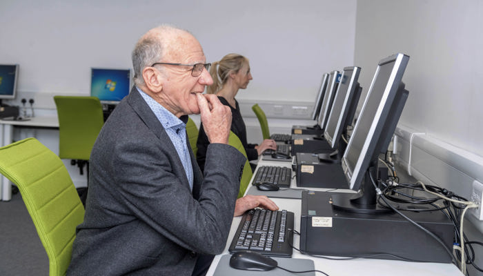 An older person wearing a grey suit looking at a computer screen with their hands on the keyboard in the library