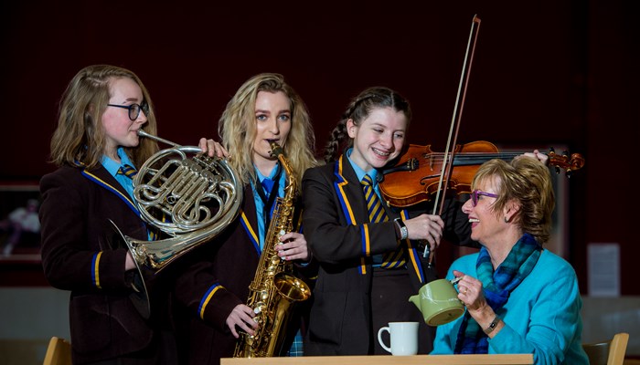 Three school girls playing instruments to a lady drinking tea. They play a violin, a trombone and a saxophone. 