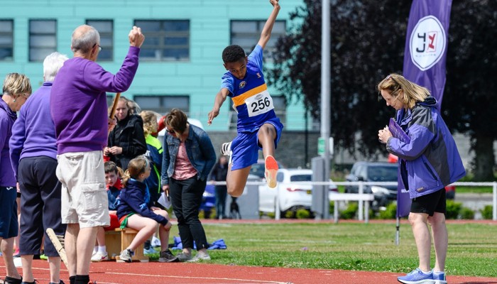 A young Shettleston Harrier athlete in mid-air during the long jump who is just about to land in the sand. Their arms are in the air and three adults, who are officiating, are watching closely.