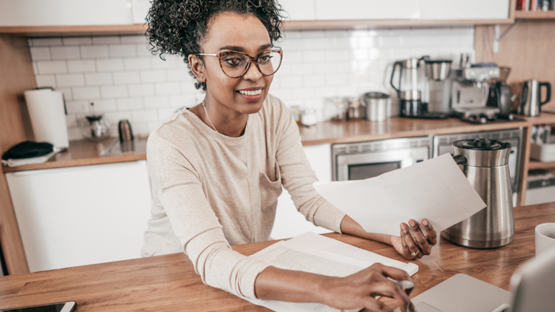 A person is working on a laptop from their kitchen. They have papers in their hand and smiling towards the screen. 