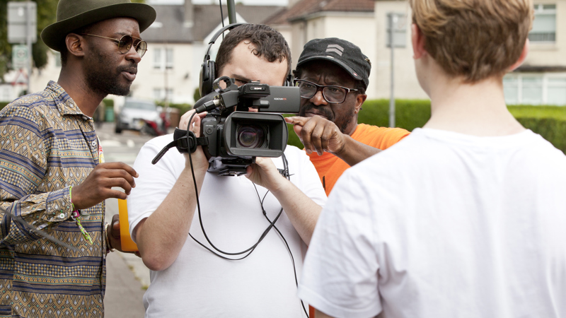 Person holding a video camera with two people on either side giving advice. The back of the person being filmed is also in view.