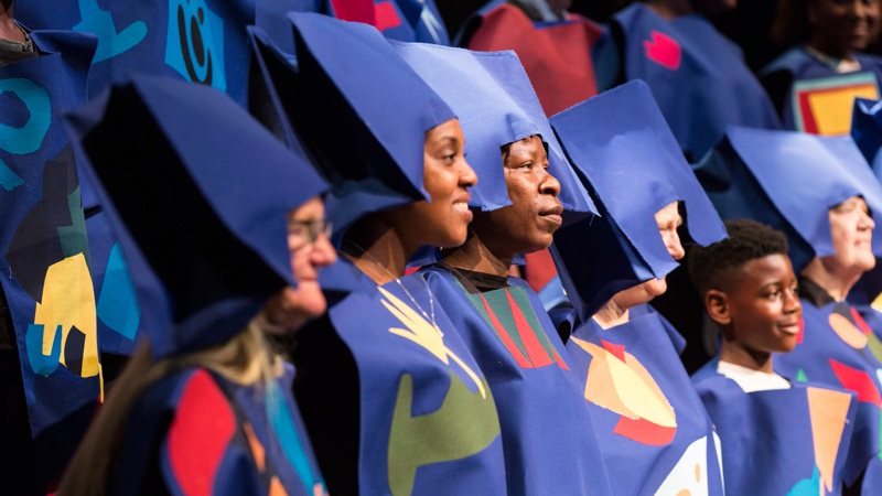 A group of mixed age people looking at a stage and smiling. Wearing blue outfits with colourful patterns and blue hats.