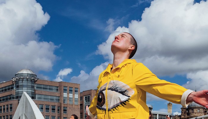 A performer dressed in a yellow jacket with a large eye on the front. They have their face to the sky and arms outstretched behind them. There are other similarly dressed performers in the background. The performance is taken place outdoors on a bridge in Glasgow and there is a blue sky and buildings in the background.