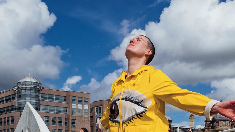 A performer dressed in a yellow jacket with a large eye on the front. They have their face to the sky and arms outstretched behind them. There are other similarly dressed performers in the background. The performance is taken place outdoors on a bridge in Glasgow and there is a blue sky and buildings in the background.