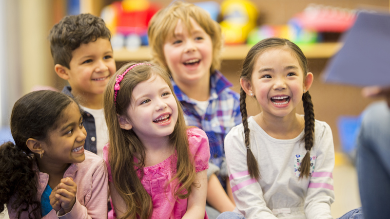 Group of children sitting on the floor looking up smiling to a person sitting out of shot on a chair holding a book. Background shows bookcases filled with brightly coloured objects