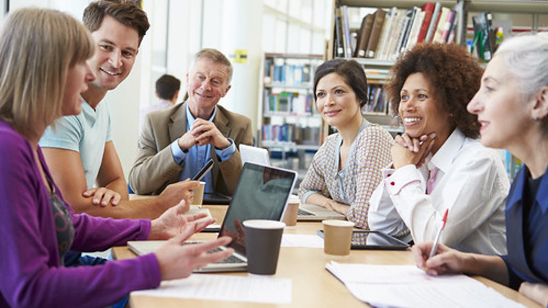 Group of adults in a library who are sitting around a table. The are all smiling and engaged in a conversation. 