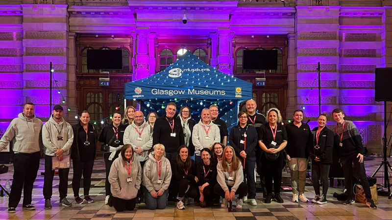 A large group of colleagues gathered for a photo in the grand marble entrance hall of Kelvingrove museum. They are in from of a blue gazebo that is branded Glasgow Museums