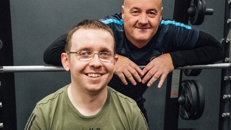 two men looking at the camera and smiling in a gym setting, one man is standing and the other is in a wheelchair, the main in the wheelchair has had his arm amputated from below the elbow. 