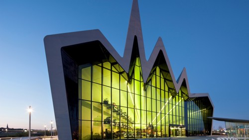 museum with roof that has 3 peaks like mountains. the front of the building is made of glass and there's a yellow reflection of the glass. the sky is blue and the shot it taken at night