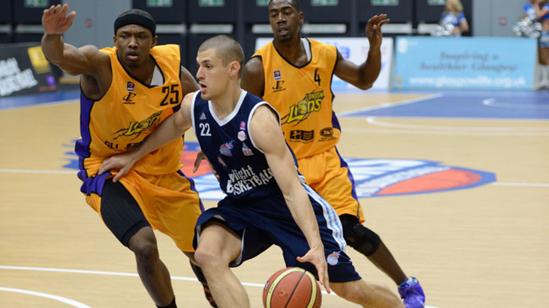 Close up of three people playing basketball indoors. The person attacking with the ball is dressed in a blue vest top and shorts and the two defenders in the opposing team are wearing yellow vest tops and shorts. They are trying to stop their opponent advancing with the ball.