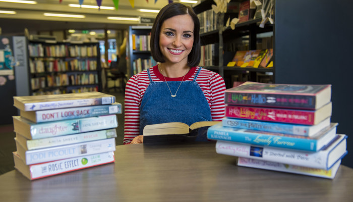 A person sitting at a table in a library between two piles of books. They are holding a book and smiling.