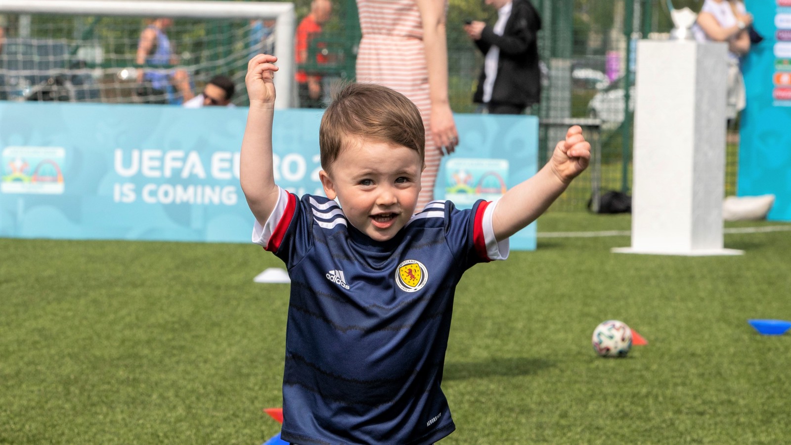 A young child smiles and raises their arms above their head while playing football