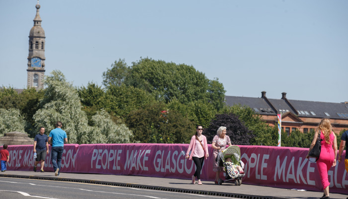 People walking across a bridge in Glasgow City Centre which has People Make Glasgow branding and signage on it.