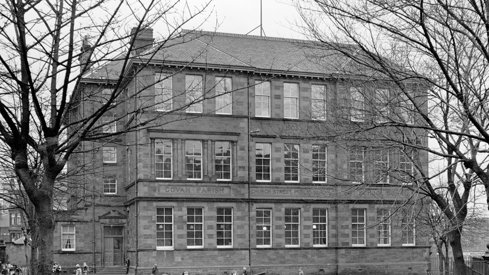 Black and white photo of Govan Parish Church Street Public School with bare tree branches coming in from the sides and children playing in the playground.