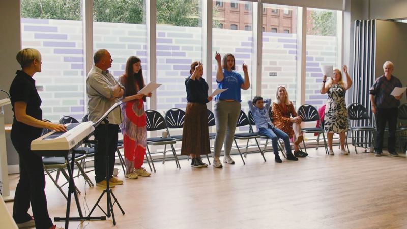 A group of people standing up during a singing for fun session, which is being led by an instructor who is standing in the corner of the room at a piano.