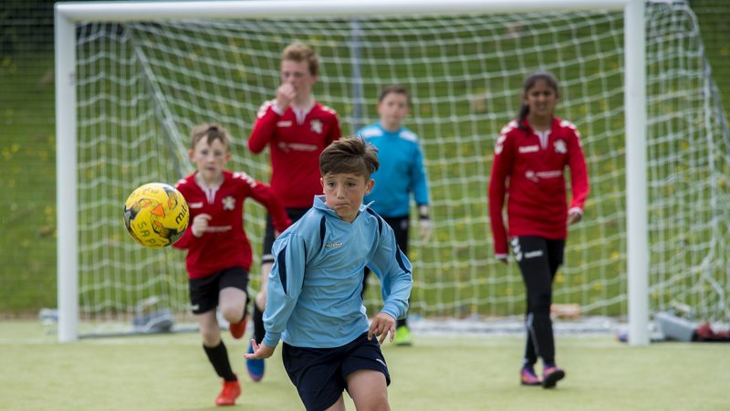 Five young football players from opposing teams. Two wearing light blue strips are attacking the ball. In the background are three players in a red strip. The game is taking place outdoors and the goal net can be seen.