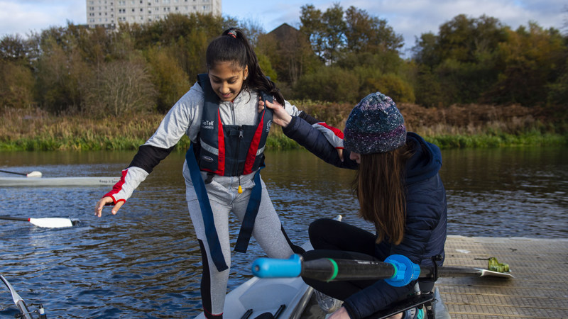 one person is crouched down on a pier beside the water while helping another as they balance and step into a canoe/kayak. They are wearing warm clothes and the one in canoe is wearing a life jacket. It is a bright day. There are oars in the background on the pier.