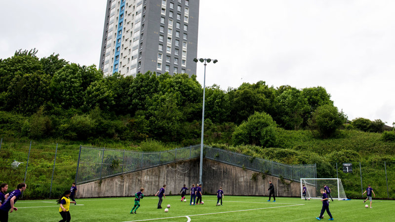 A high rise residential building is in the background. In front is a football pitch with young players training. There are hedges around and quite a few footballs on the pitch.