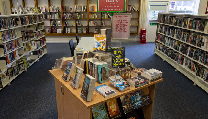 A view of the reading area of Baillieston library. There is a wooden bookstand in the middle of the room which is surrounded by more bookshelves.