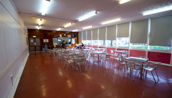 large room with multiple tables are chairs places around the room. the floor is dark orange and the walls are white. there are windows lining the right hand side of the room