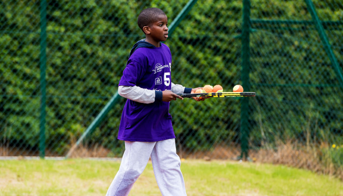 A young person playing tennis, they are holding a racket in their right hand which has three brightly coloured tennis balls on it.