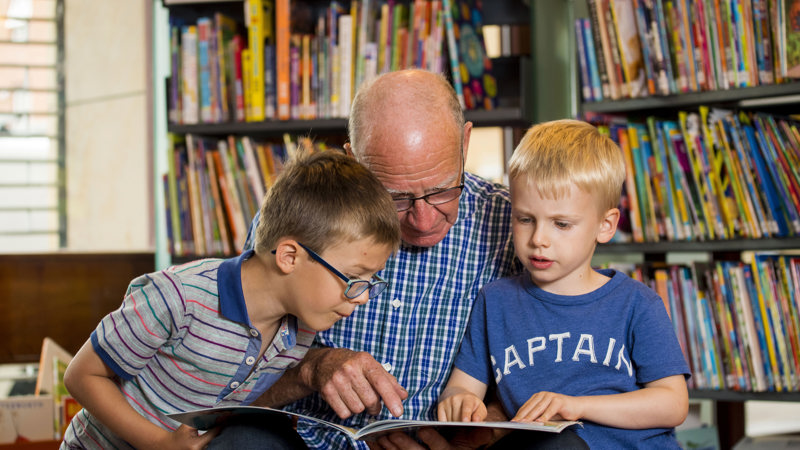 An adult is reading and pointing at a book with a child on each side. They are in a library and there are shelves of books in the background.