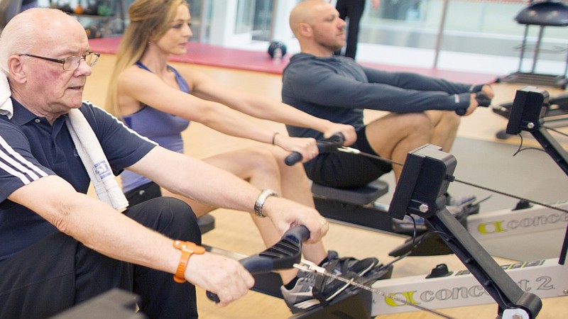 three people of varying ages all working out on rowing machines within the gym