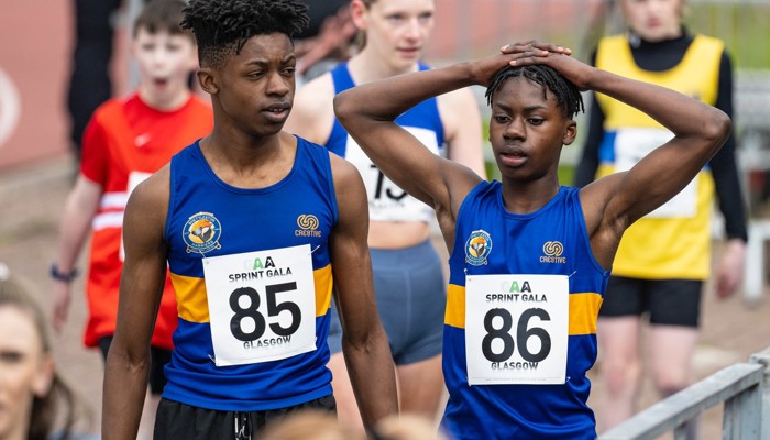 Two young Shettleston Harrier athletes at a competition, both are wearing club vest which are blue and yellow. One of the young athletes has their hands on their head.