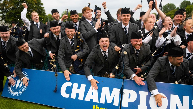 A large pipe band, all dressed in kilts, are celebrating their win outside behind a banner that reads Champions, trees and blue skies are visible in the background.