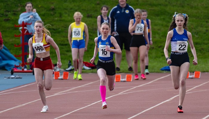 Three young athletes racing during an athletics sprint event. They are wearing different club colours and are in separate lanes on the track.