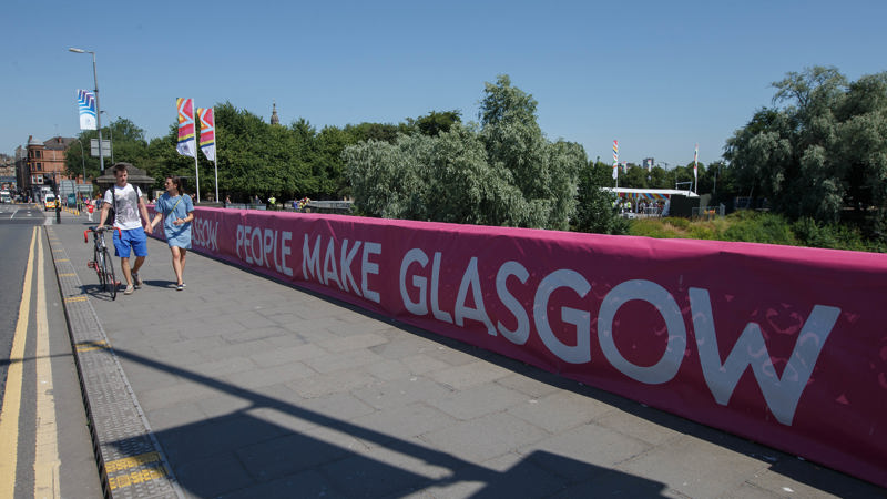 The People Make Glasgow branding on a bridge across the River Clyde in Glasgow city centre. A couple are walking across the bridge and one of them has a bike.