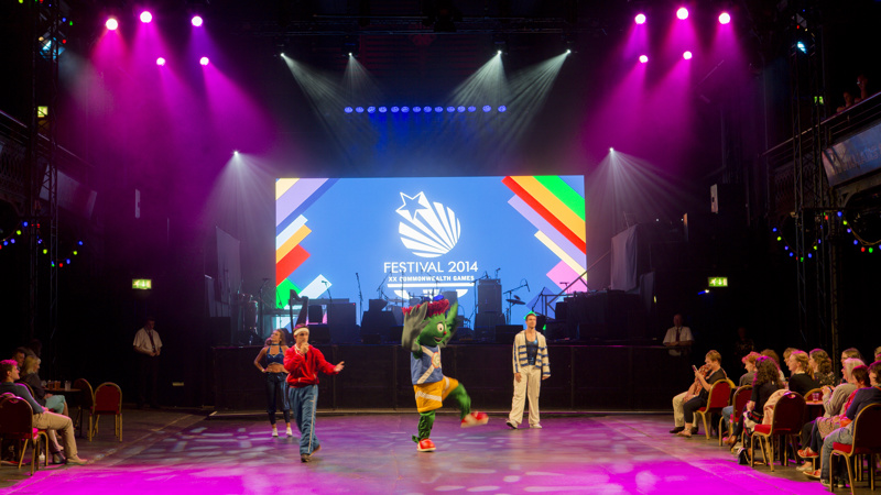 Clyde the Thistle mascot dancing with other performers at the Old Fruitmarket in Glasgow in front of a large screen which say Festival 2014.