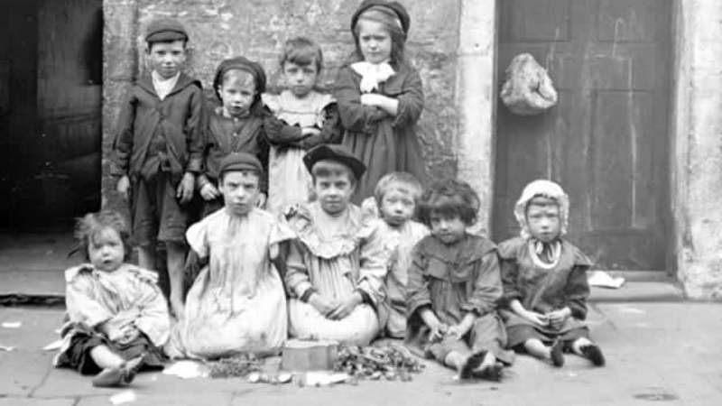 Black and white photo showing a group of 10 children of different ages in two rows, 4 children are standing in the back row whilst the rest are sitting in front row, there is a building and door behind them