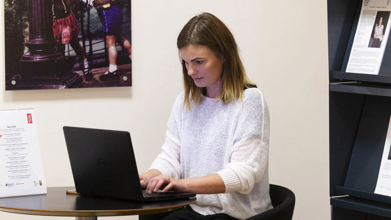 A women sitting at a small desk typing on a laptop with a bookcase behind her with information resources on the shelves