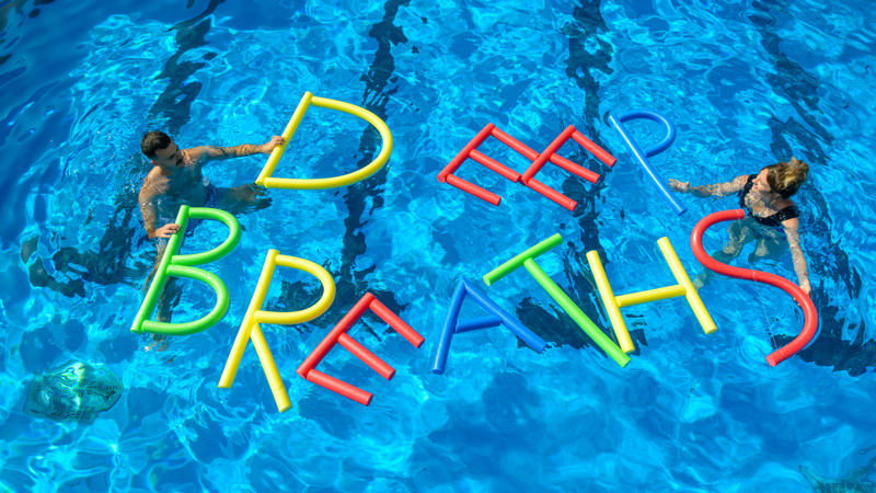 Two people are in a pool either side of floats which spell out 'Deep Breaths' 