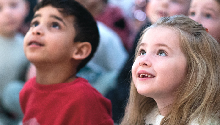 Two primary age children are sitting on the floor looking up at a performance on stage and smiling. There are other children sitting in the background.