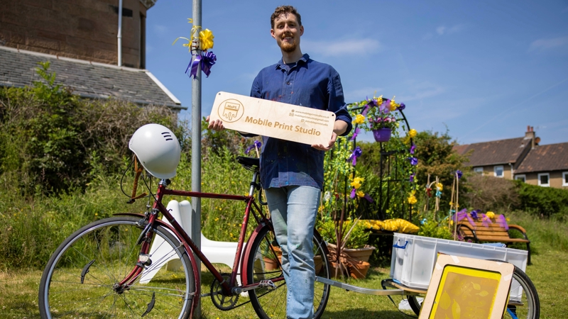 A man is pictured in front of his bike and some craft equipment. he's holding a sign that reads 'mobile print studio'