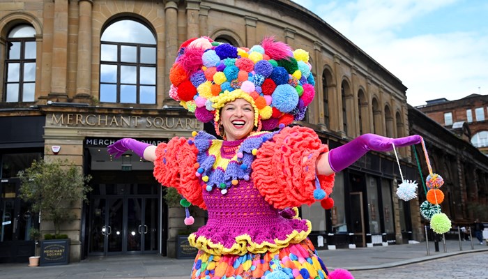 A performer in a brightly coloured outfit made of pom-poms stands in front of a 1700s building. A sign for Merchant Square can be seen behind the performer.