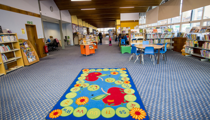 A bright, spacious children's area with a blue checked carpet, large windows along the right hand side and a wood pannelled ceiling. There is a round wooden table with blue chairs and a colourful children's rug in the centre of the room.